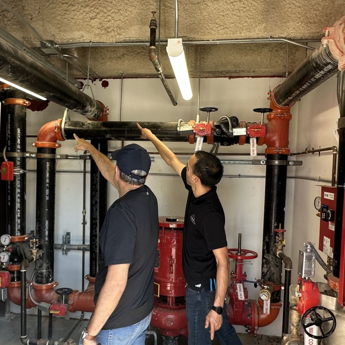 Two Paragon technicians inspecting the overhead feed main and valves in a commercial fire sprinkler riser room