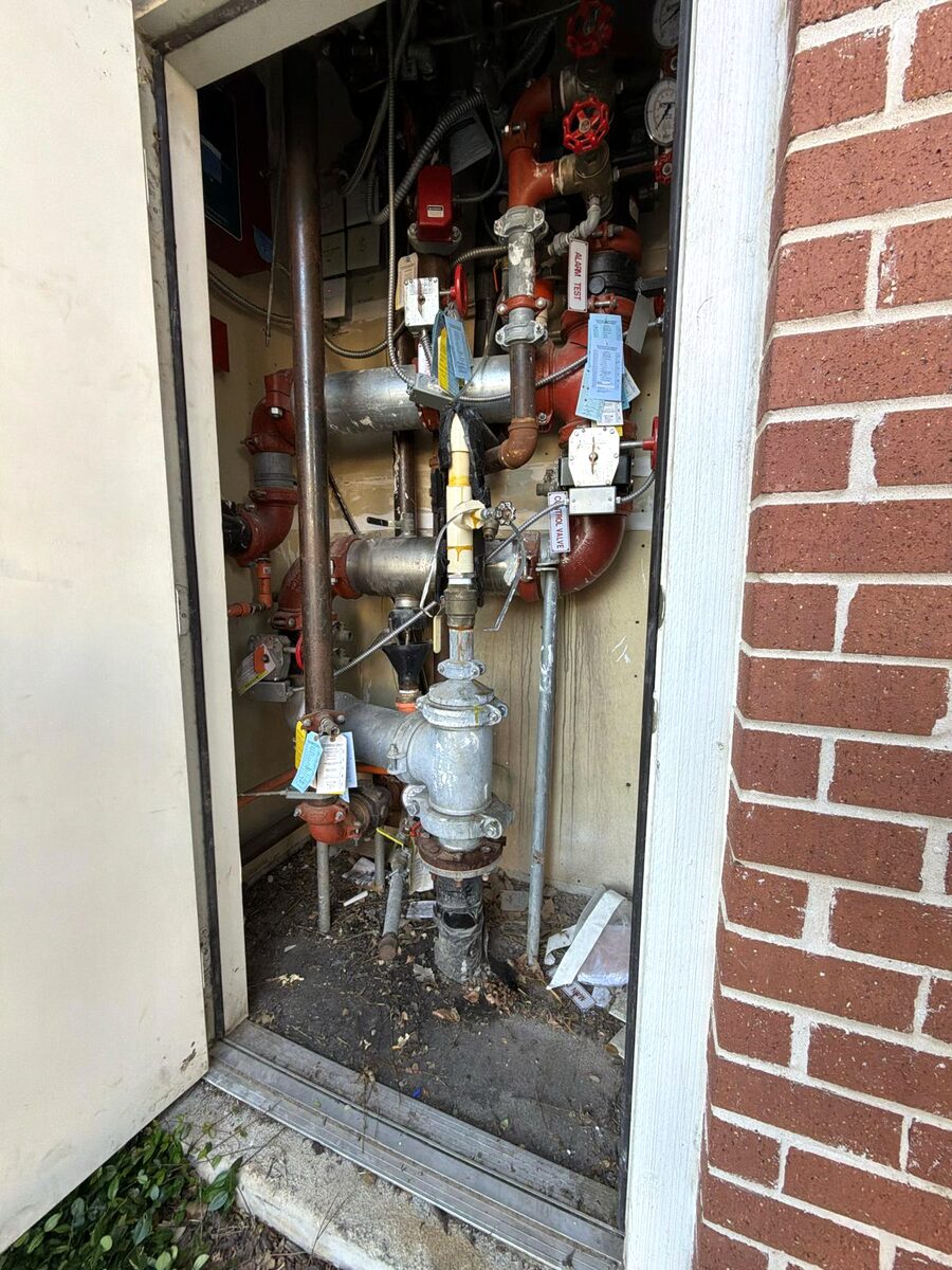 The cramped exterior riser closet the new compressor had to be fitted into — sprinkler riser, backflow, and valving packed into a narrow space